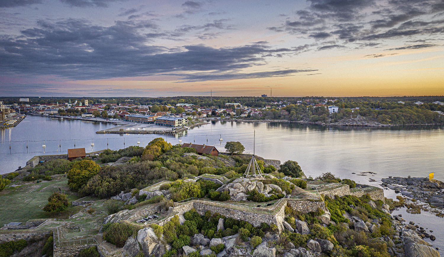 Vy av en hamn och stad vid solnedgång, med stenar och grönska i förgrunden.