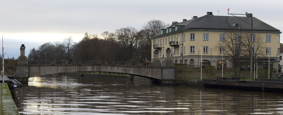 Flod med stenbro och byggnad i bakgrunden, omgiven av träd under molnig himmel.