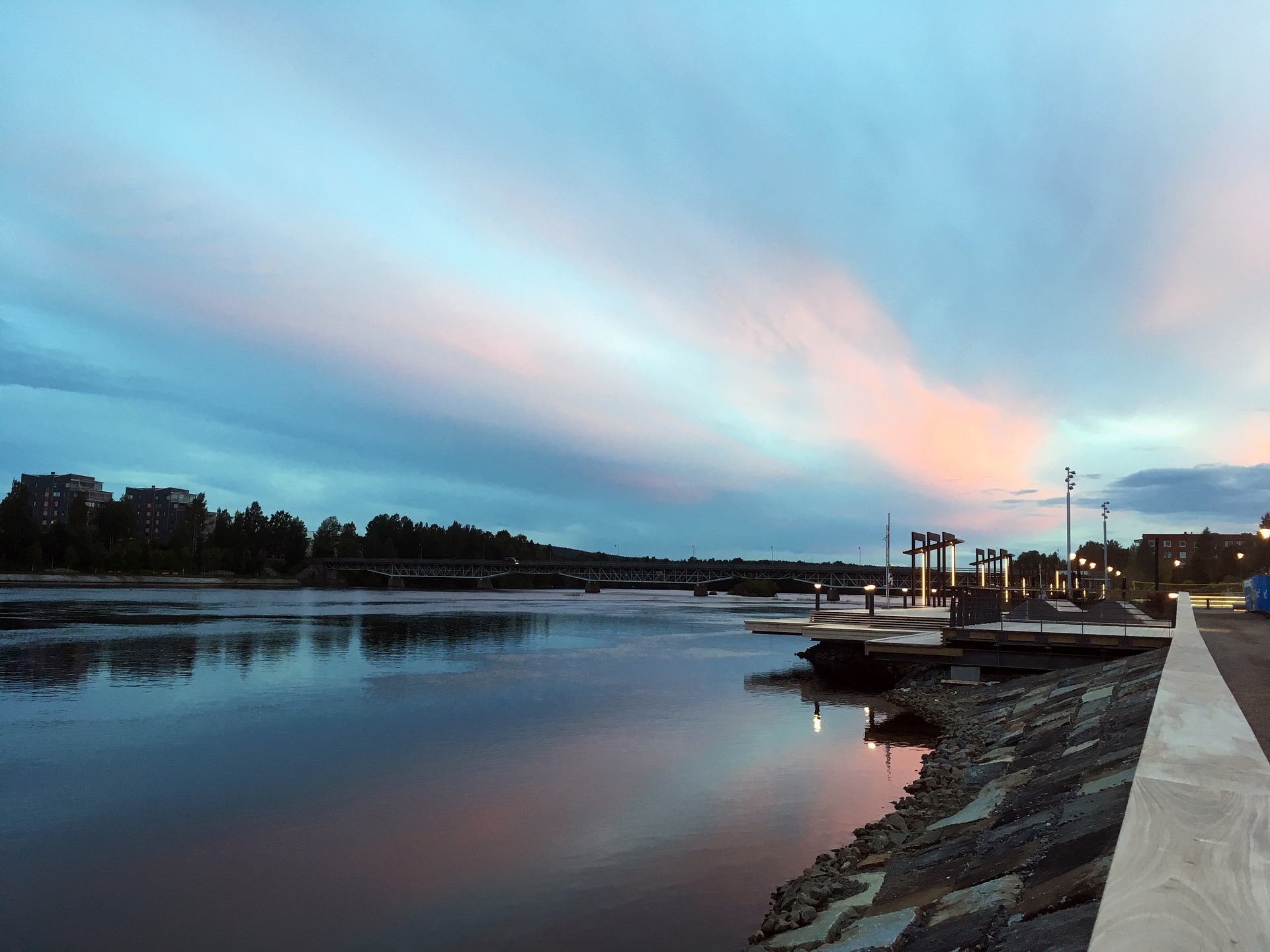 Flodlandskap i solnedgång, med himmel i rosa och blåtoner över vattnet och staden i bakgrunden.