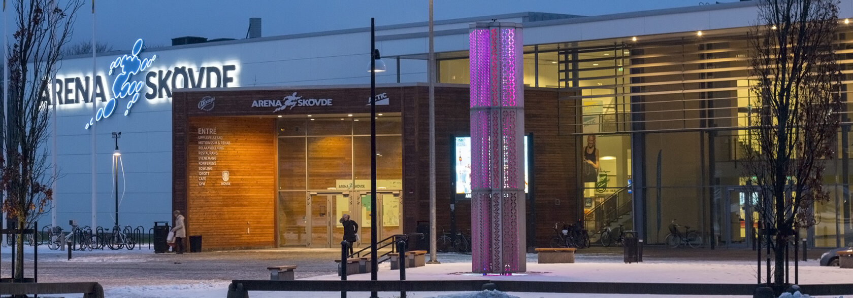Byggnad exteriör, skylt med texten "Arena Skövde" upplyst. Snöig mark, rosa belyst stolpe framträdande.