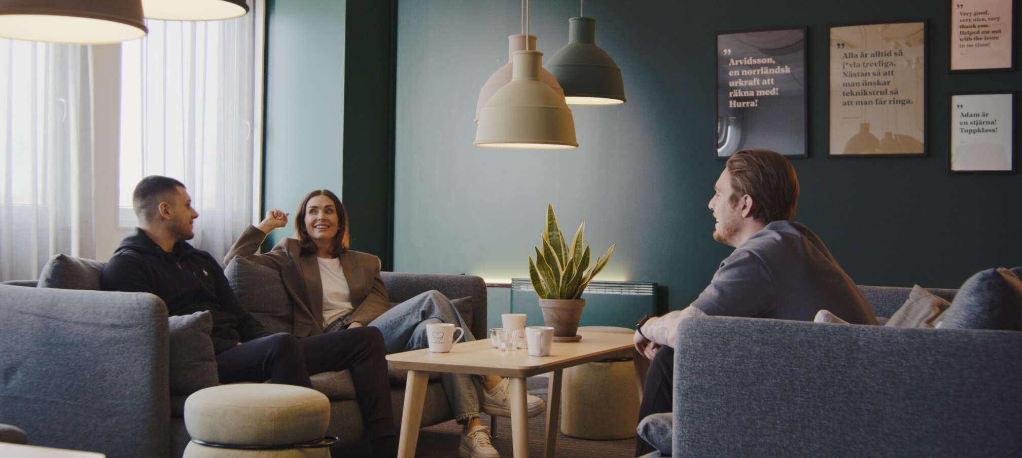 Tre personer sitter och pratar avslappnat i en soffa med väggcitat och en krukväxt i närheten.