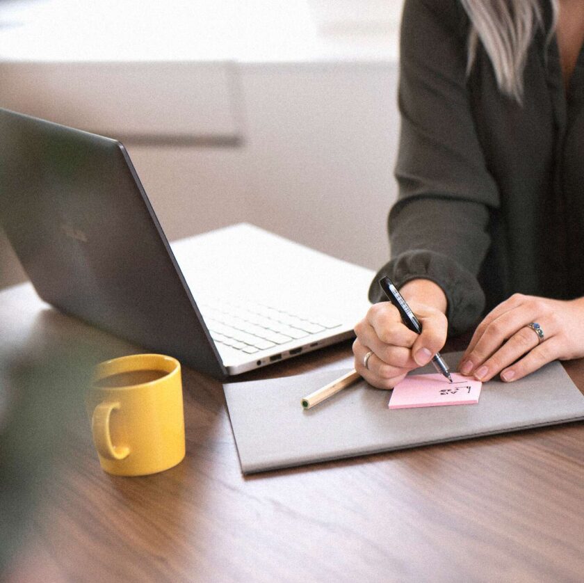 Person skriver på en rosa post-it vid ett skrivbord med laptop och gul kopp.