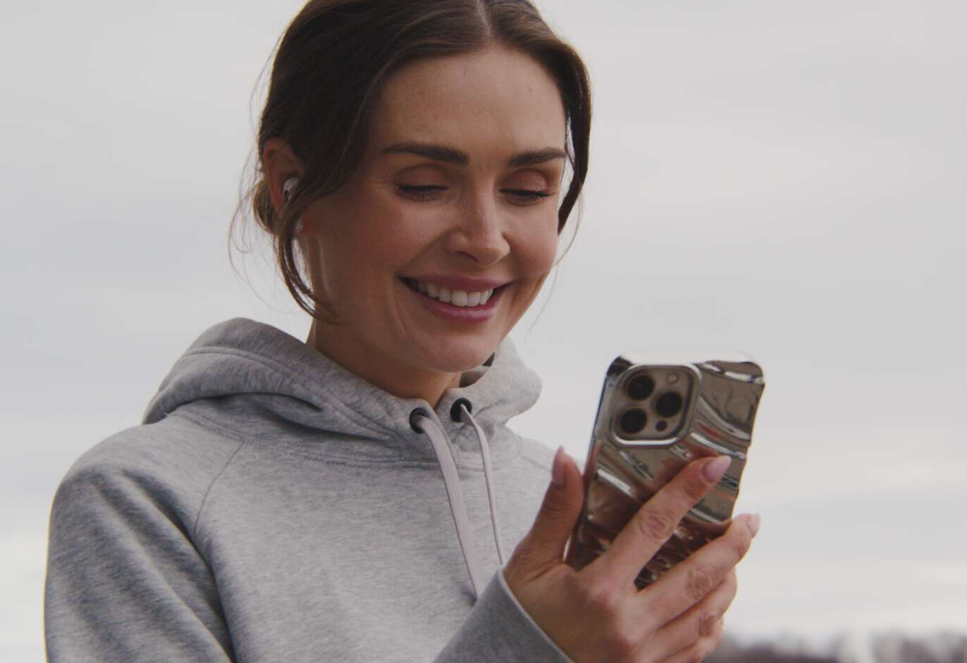 En person i grå hoodie ler medan hen tittar på en mobiltelefon utomhus.