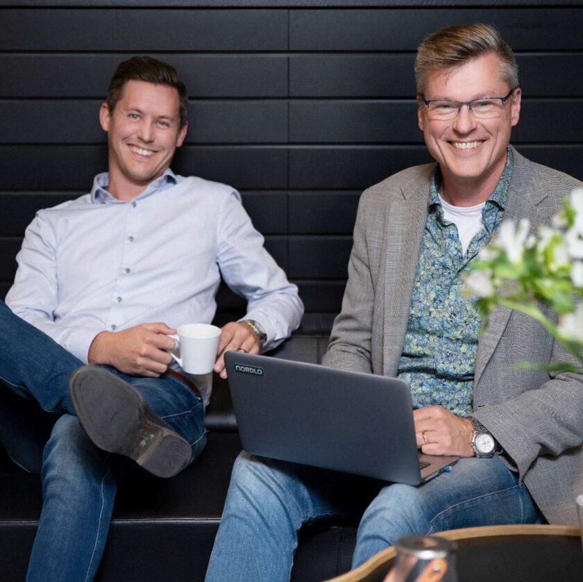 Två leende män sitter i en soffa, en med en kopp och den andra med en bärbar dator.