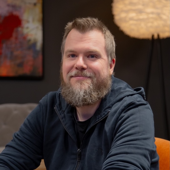 Skäggig man i mörk tröja, sitter inomhus med en färgglad tavla och en rund lampa i bakgrunden.