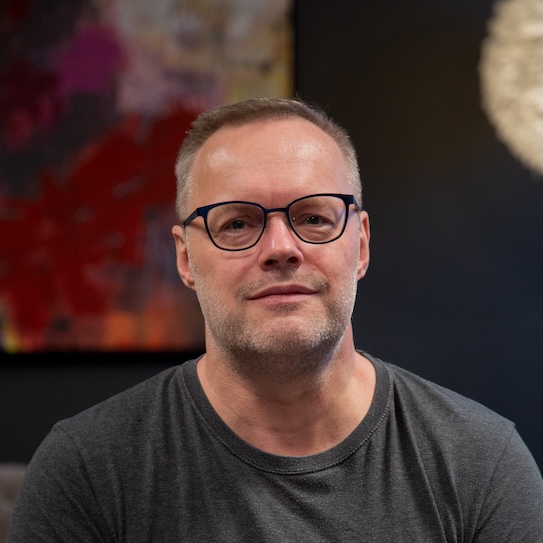 En man med glasögon och grå t-shirt står framför en färgglad tavla och mörk bakgrund.
