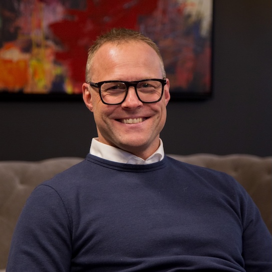 En person med glasögon och blå tröja sitter leende på en soffa, med en färgglad tavla i bakgrunden.