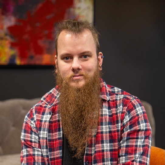 Man med långt skägg och rutig skjorta sitter framför en färgglad tavla.