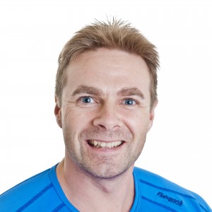 En smilende person i blå t-skjorte foran en hvit bakgrunn.