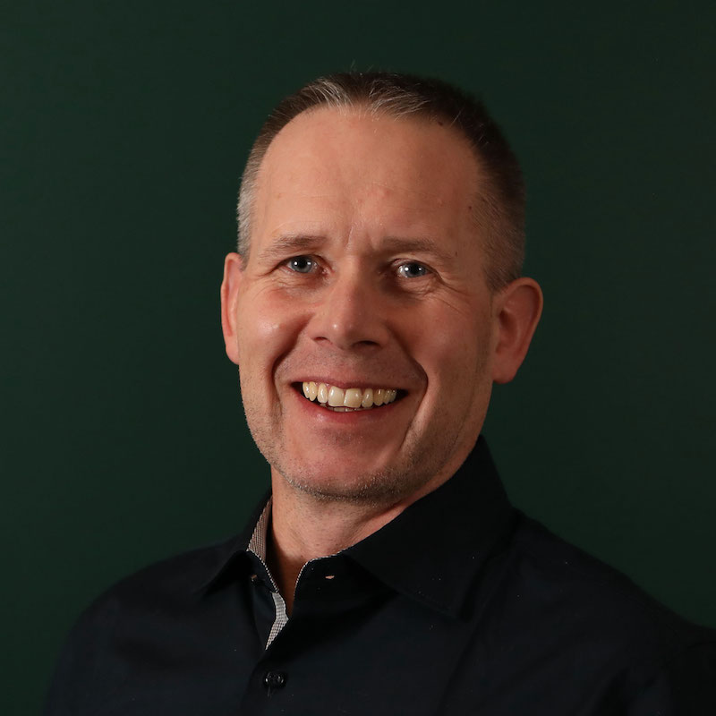 En smilende mann med kort hår i en mørk skjorte, grønn bakgrunn.