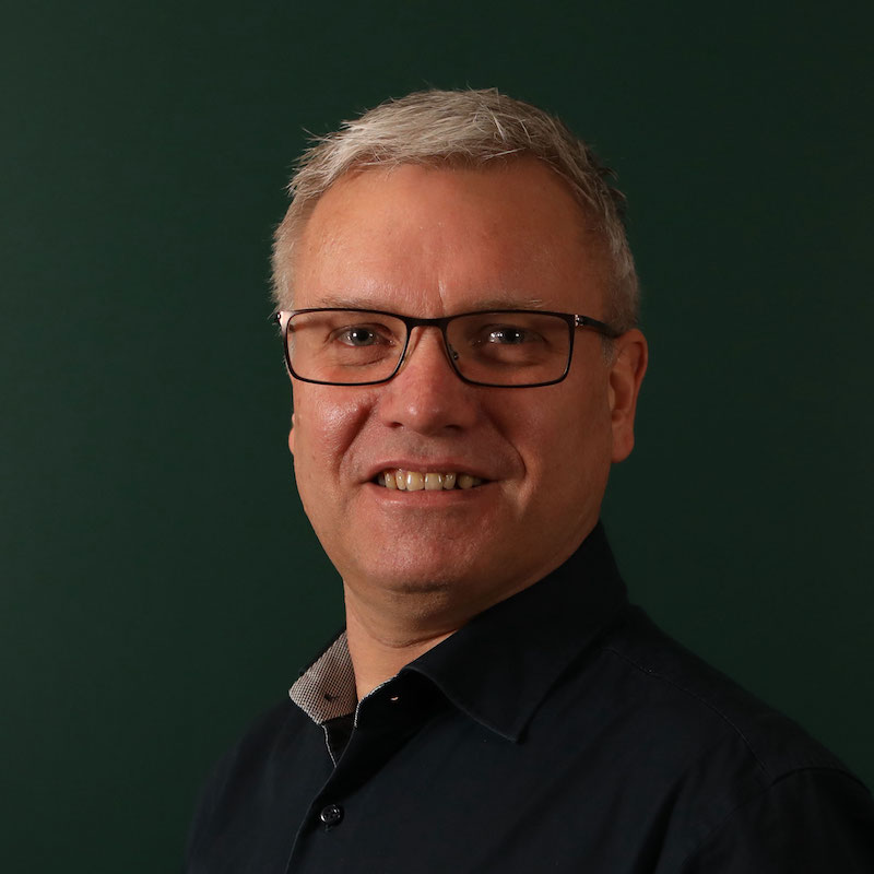 En mann med briller og grått hår smiler mot kameraet, iført en mørk skjorte.