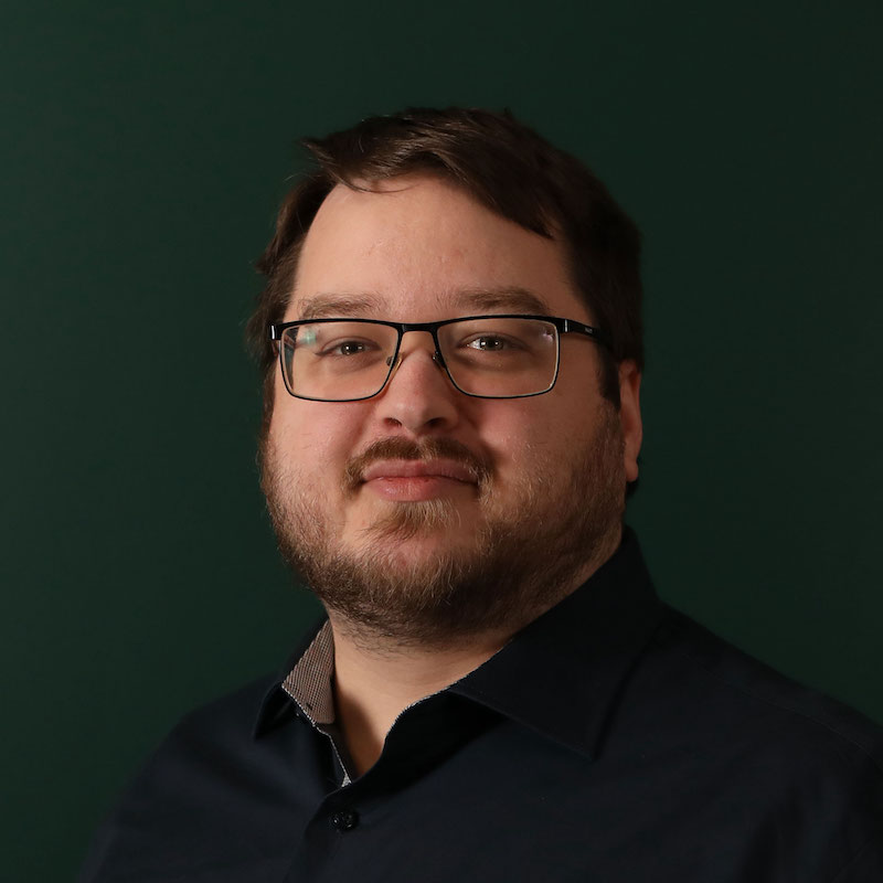 En mann med briller og skjegg mot en mørk grønn bakgrunn.