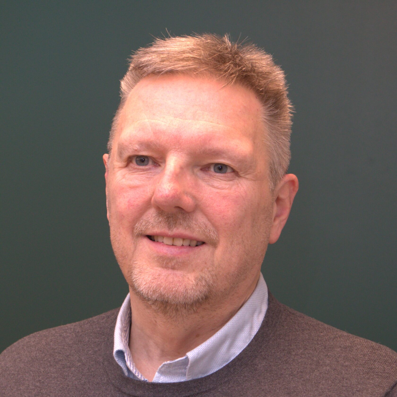 Mann med kort hår og skjeggstubb, iført en genser over en skjorte, mot en mørk bakgrunn.