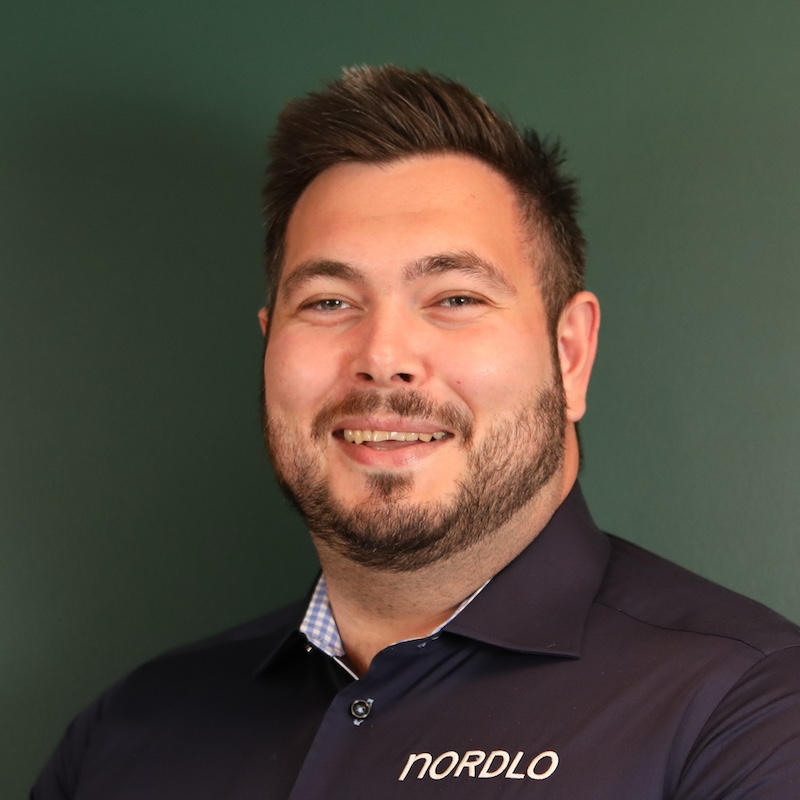 En smilende mann med skjegg i mørk skjorte foran en grønn vegg.