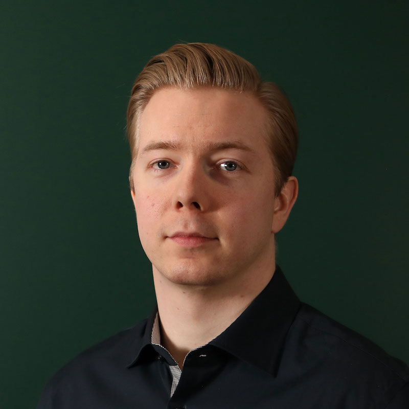 En person i mørk skjorte ser mot kameraet med en nøytral ansiktsuttrykk og en mørk grønn bakgrunn.