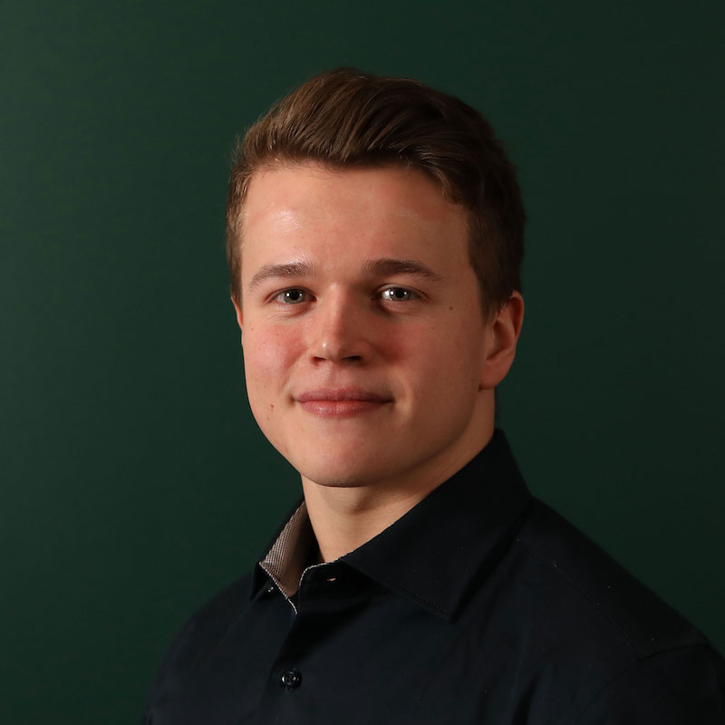 En mann med kort, brunt hår iført en mørk skjorte smiler foran en grønn bakgrunn.