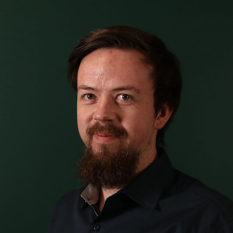 En mann med skjegg og mørkt hår foran en grønn bakgrunn.