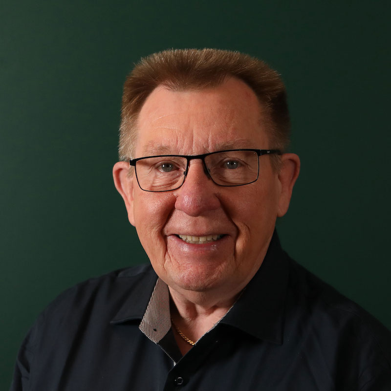 Eldre mann med briller smiler foran en mørk grønn bakgrunn.