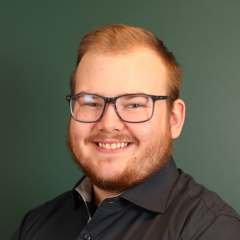 En smilende person med briller og skjegg, kledd i en mørk skjorte, mot en grønn bakgrunn.