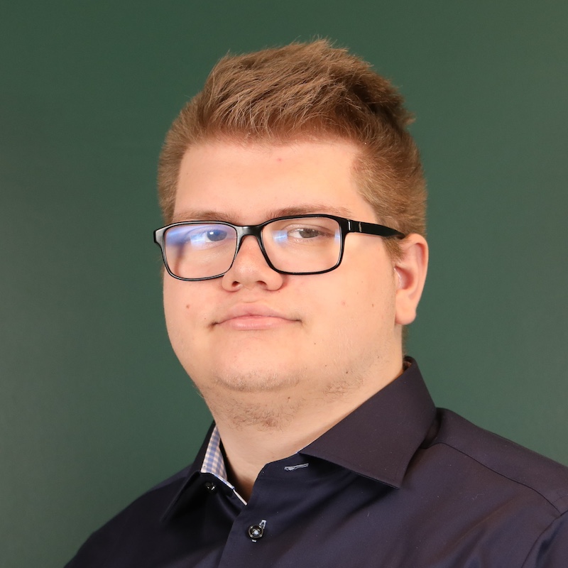 Unge mann med briller og mørk skjorte foran en grønn bakgrunn.