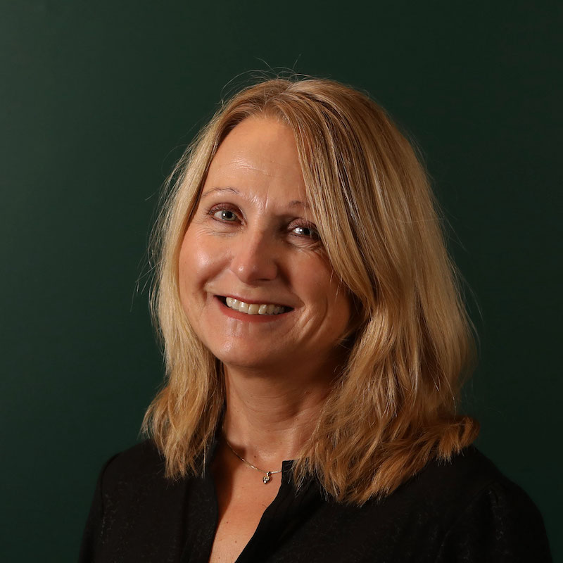 En kvinne med blondt hår smiler mot kameraet med en mørkegrønn bakgrunn.