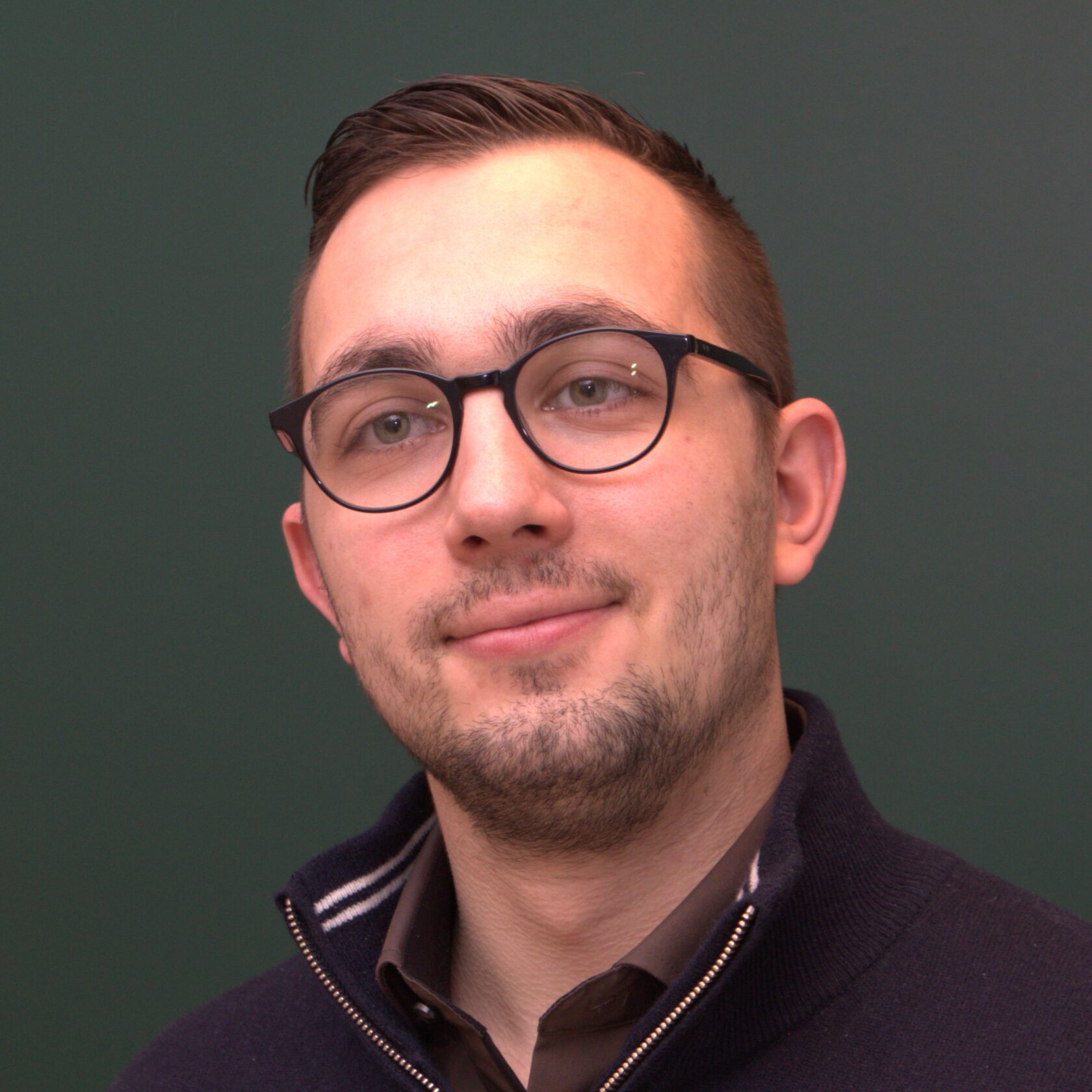 Mann med briller og mørkt hår i en mørk jakke foran en grønn bakgrunn.