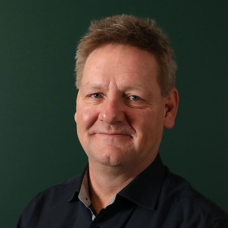 En person med kort hår og mørk skjorte smiler mot kameraet, bakgrunn grønn.