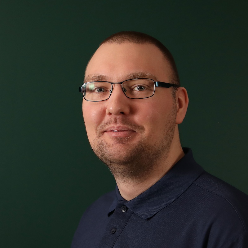 En mann med briller og mørk polo står foran en mørk bakgrunn.