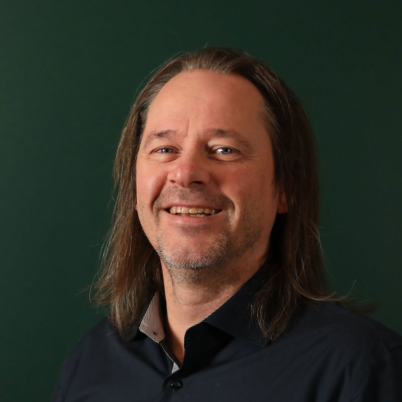 En smilende mann med langt hår og skjegg, iført en mørk skjorte, mot en mørk grønn bakgrunn.