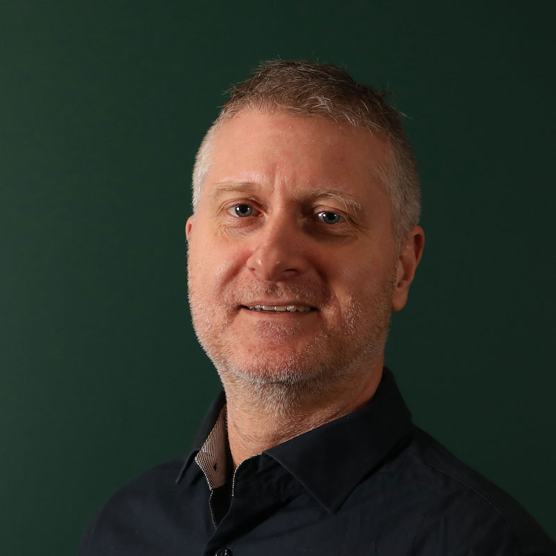 En mann med kort hår og mørk blå skjorte smiler foran en mørk grønn bakgrunn.