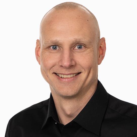 Man med rakad huvud och svart skjorta ler mot kameran.