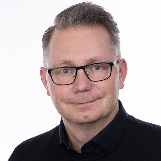 En man med glasögon, kort hår och svart tröja ler mot kameran. Bakgrunden är vit.