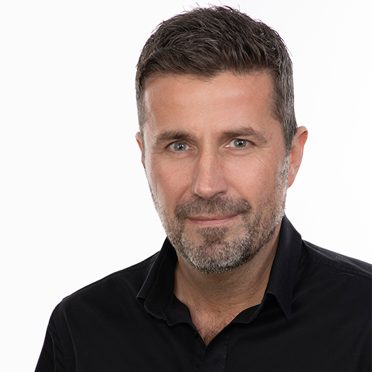 Man med kort hår och skägg, bär en svart skjorta, ser mot kameran.