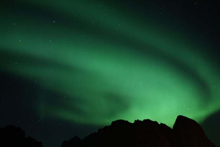 Nordlys danser grønt over mørke fjelltopper under en klar nattehimmel med stjerner.